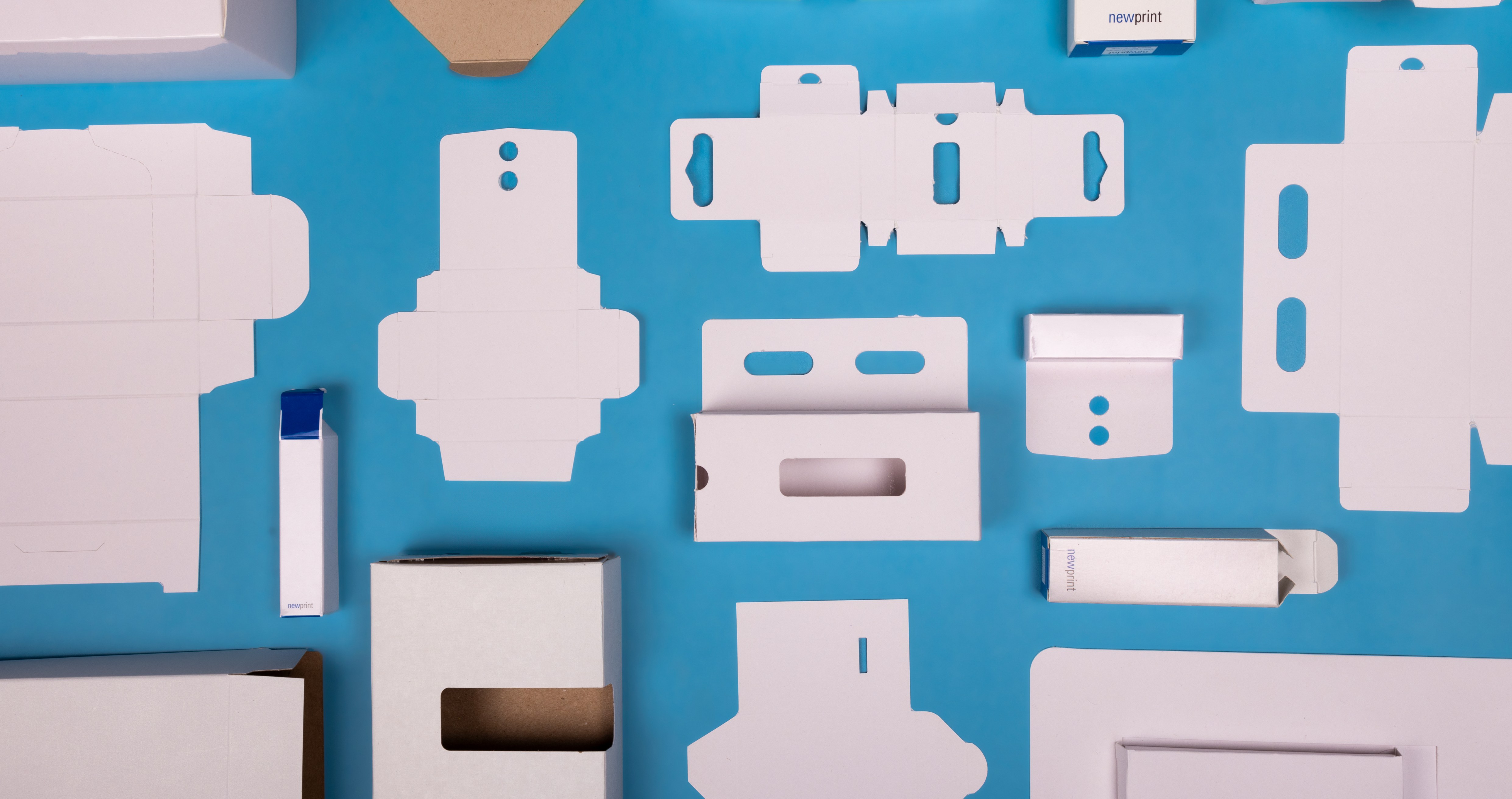 Various cardboard boxes and other items are on a blue surface, representing packaging samples and the prototyping workflow.